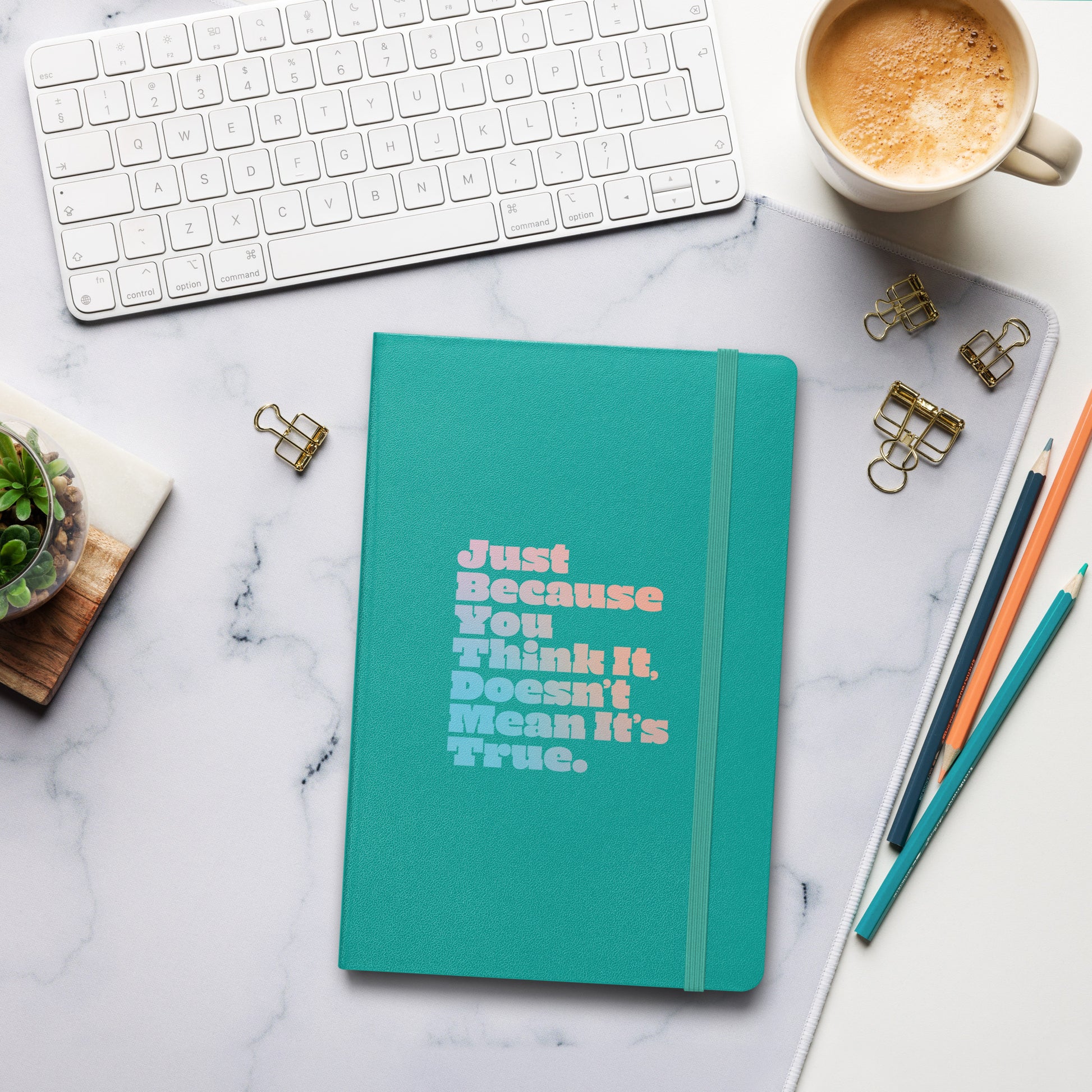 Just Because You Think It, Doesn't Mean It's True type on turquoise notebook, front view on work desk with accessories and coffee cup nearby